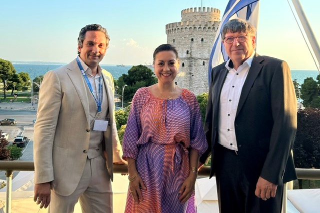 Drei Personen stehen lächelnd vor dem Weißen Turm in Thessaloniki unter griechischer Flagge.